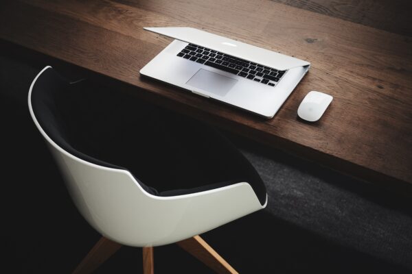 A sleek workspace featuring a partially open MacBook, Apple Magic Mouse, and a modern white chair at a wooden desk