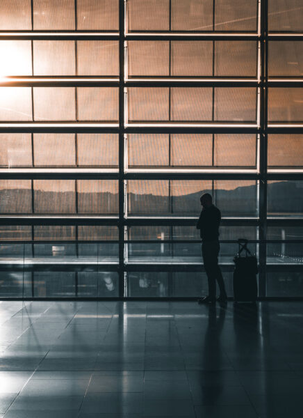 A person stands in front of large, sunlit airport windows at sunset with a suitcase beside them.