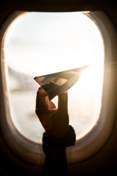 A hand holding a paper airplane in front of an airplane window, with bright sunlight streaming through