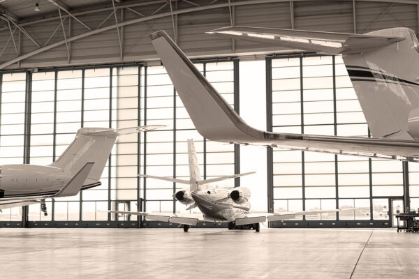 Three private jets parked in a spacious, well-lit hangar with large windows, showing the tails and engines of the aircraft.