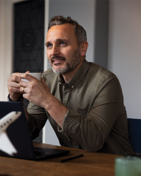 A man in a green shirt holding a coffee cup, smiling while sitting at a table with a laptop