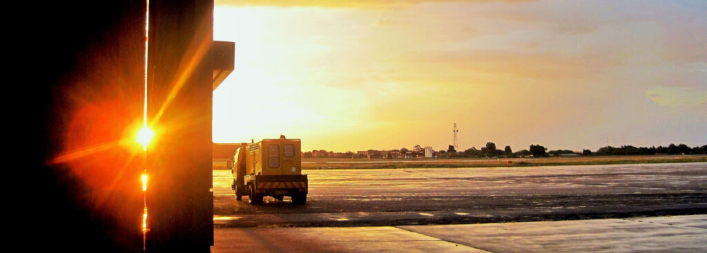Image of a yellow truck driving out onto an airport tarmac with light coming through a column