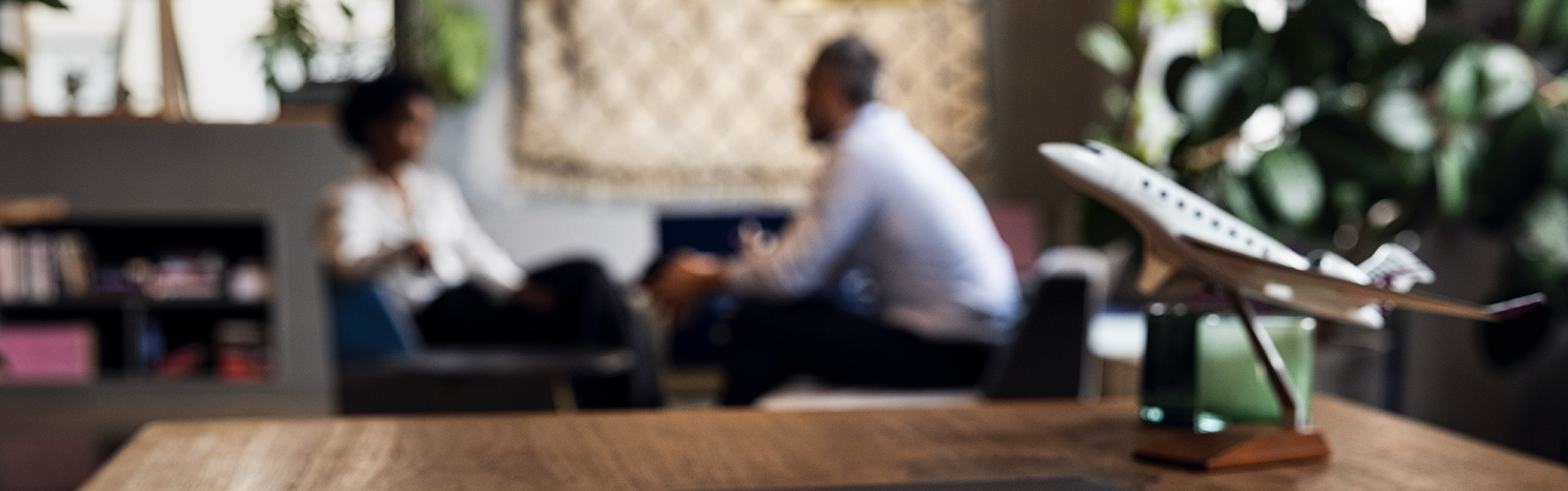 Image of wooden desk with a black ipad and model airplane in focus while a meeting is taking place in the blurred background