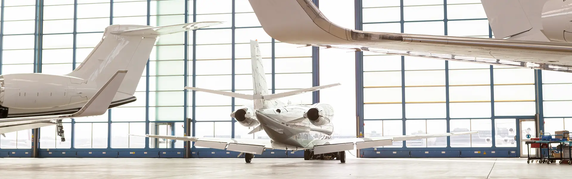 Three private jets parked in a spacious, well-lit hangar with large windows, showing the tails and engines of the aircraft.