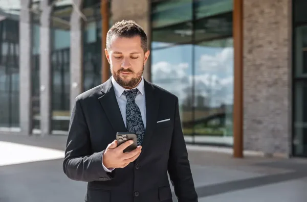 Man in a suit holding a phone, standing outdoors with a modern building in the background.