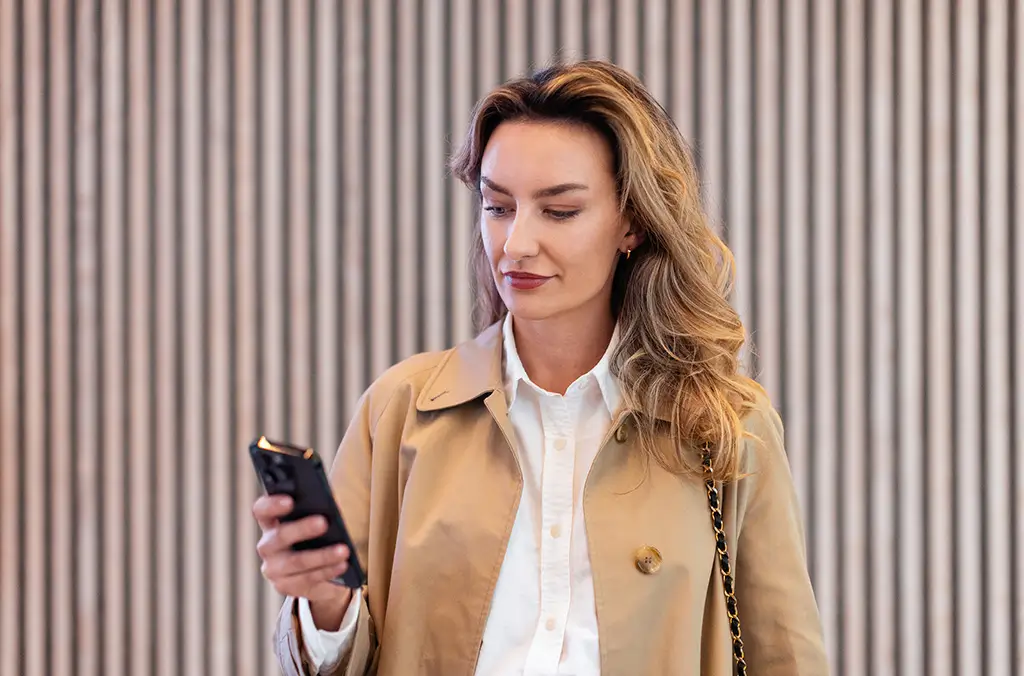 A woman in a white shirt and a beige coat looks at her phone while standing in front of a striped background