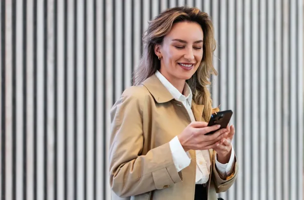 A woman in a white shirt and a beige coat smiling at her phone while standing in front of a striped background