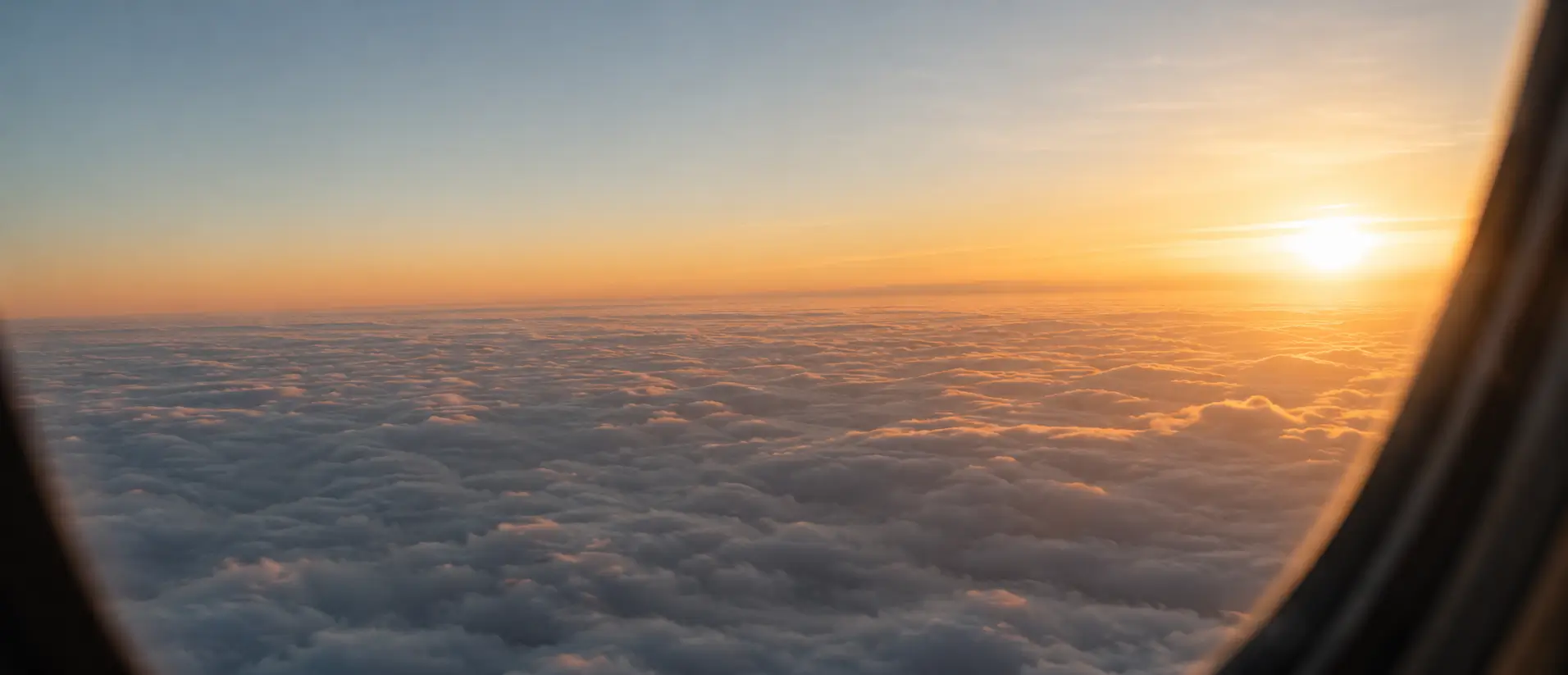 Hero image looking out an airplane window at a sunset above the clouds.